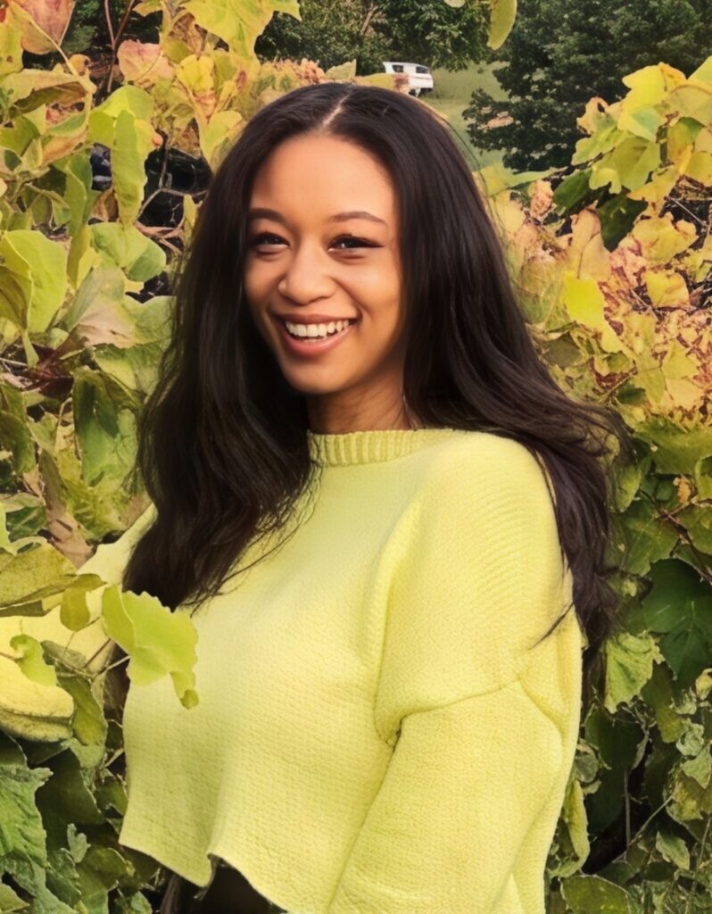 Jasmine Scott A woman with long, dark hair wearing a light green sweater, standing outdoors among green and yellow foliage, smiling and looking slightly to her right.