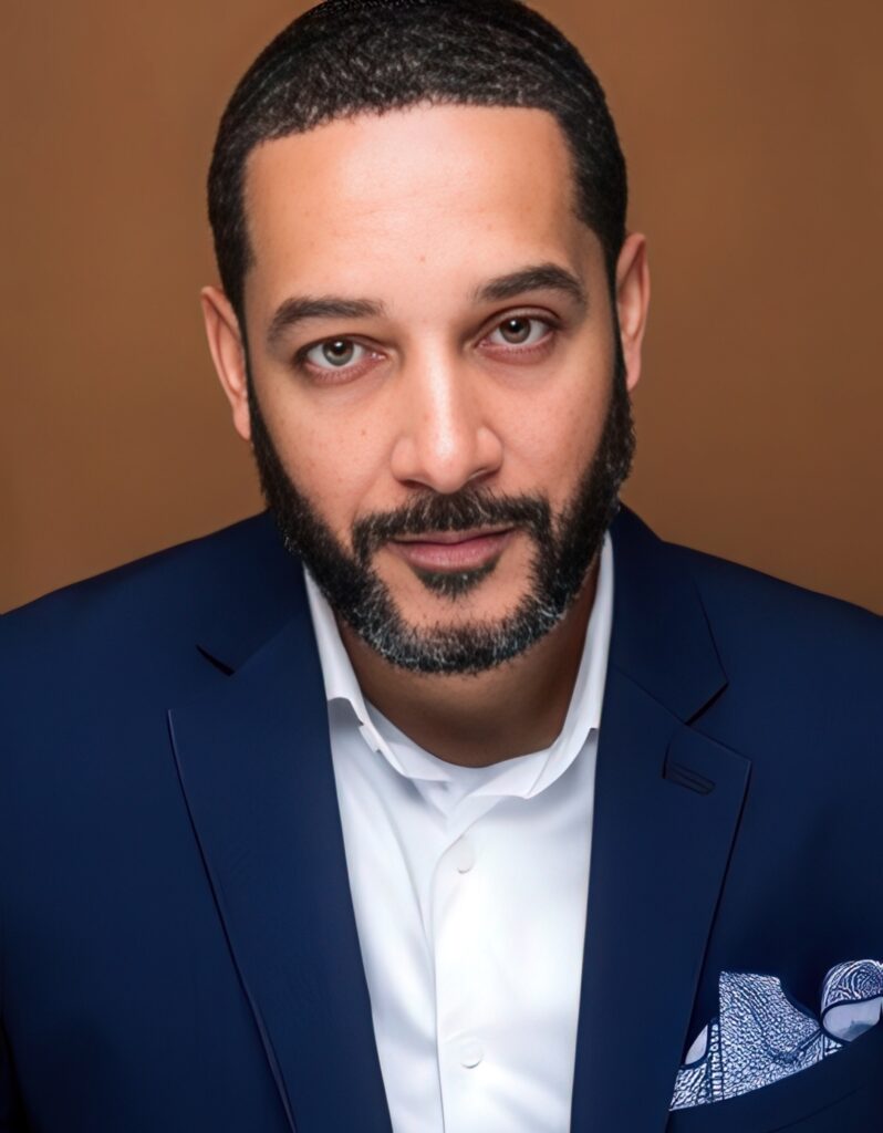 Shawn Rux A man with a short beard and neatly trimmed hair wearing a navy blue suit jacket and white shirt, looking directly at the camera with a neutral expression, against a plain brown background.
